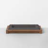 A modern rectangular pet bed with a wooden frame and dark gray cushioned interior, placed on a light gray floor against a plain white wall.