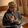 A man wearing glasses, a tan cap, and a blue hoodie sits on the edge of a bed, holding a gray bowl of colorful cereal and a spoon, bathed in warm sunlight. Text reads: Thuma • Heller Max Breakfast Set.