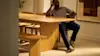 A man sits thoughtfully at a large wooden dining table in a softly lit, modern room with beige carpeting and minimal decor.