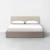 A modern bed with a beige upholstered headboard, two white pillows, and white bedding. The bed frame is made of light wood and sits on a light gray floor against a plain white wall.