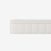 A close-up side view of a white mattress with a quilted, textured surface and stitched vertical panels. The background is plain and white.