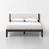 A modern wooden bed frame with a light upholstered headboard, white bedding, and two white pillows, set against a plain white wall and gray floor.