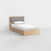 A single wooden bed with a grey upholstered headboard, white bedding, and a pillow, placed in a minimal, white-walled room with a light-colored floor.