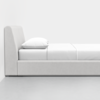 A minimalistic, modern bed with a padded headboard and base upholstered in light gray fabric, featuring crisp white bedding and a single pillow, placed against a plain white wall in a bright room.