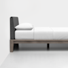 A minimalist bed with a gray upholstered headboard, white bedding, and a wooden frame is shown against a plain white wall and floor, viewed from the side.