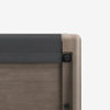 Close-up of the corner of a wooden bed frame with a dark gray upholstered headboard, showing the texture of the wood and fabric against a plain white background.