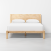 A wooden bed frame with a simple headboard and legs, made of light-colored wood, is shown with a white mattress, white sheets, and two white pillows, set against a plain white wall and floor.