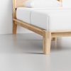A close-up view of a wooden bed frame with a white mattress and pillow, set on a light gray floor against a white wall.