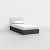 A single bed with a dark wooden frame, white mattress, white pillow, and a light-colored upholstered headboard, set against a plain white wall and floor.