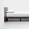 A minimalist bed with a gray upholstered headboard, white bedding, and a single pillow, set against a plain white wall and floor.