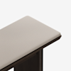 A close-up view of the corner of a bench with a light beige upholstered cushion and a dark wooden frame set against a white background.