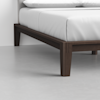 Close-up view of the corner of a dark wooden bed frame with a white mattress and bedding, set on a light gray floor against a white wall.