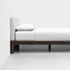 A minimalist bed with a padded light gray headboard, white bedding, and a dark wooden frame, set against a plain white wall and white floor.