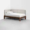 A minimalist wooden daybed with beige upholstered back and side panels, a white mattress, and a single white pillow sits in the corner of a plain, light gray room.