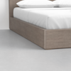 A close-up view of the corner of a wooden platform bed with white bedding and pillows, positioned on a smooth, light gray floor against a plain white wall.
