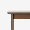 A wooden bench with a rectangular seat cushion upholstered in light beige fabric, set against a plain white background. Only the left side of the bench is visible in the image.
