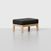 A minimalist ottoman with a light wooden frame and a rectangular, black upholstered cushion, placed on a smooth, light gray floor against a plain white background.