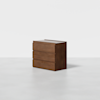 A modern, three-drawer wooden dresser with a smooth, dark brown finish, standing alone on a light grey floor against a plain white background.