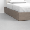 A close-up of a modern bed with a light wood platform frame, white bedding, and a white pillow, placed on a light gray concrete floor.
