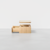 A light wooden box with a removable lid is shown. One side has an open drawer, and the lid is hovering above the box, revealing compartments inside. The background is a plain, light gray surface and wall.