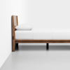 A minimalist wooden bed frame with a headboard, white bedding, and a single pillow, positioned against a white wall on a light gray floor.