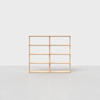A wooden shelving unit with eight open rectangular compartments is centered on a plain, light gray floor and background. The shelves are simple and minimalist in design.