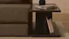 A modern wooden side table next to a brown sofa, holding a stack of magazines and a black object on top, with a few books stored on the lower shelf, placed on a light-colored rug.