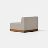 A modern armless chair with light beige upholstery sits on a low, rectangular wooden base. The chair is positioned at an angle on a light gray floor, against a plain white background.