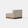A modern armless chair with light beige upholstery sits on a low, rectangular wooden base. The chair is positioned at an angle on a light gray floor, against a plain white background.