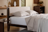 A neatly made bed with a beige upholstered headboard, two pillows, and a light comforter stands in a modern, minimalist bedroom with wooden furniture and neutral decor accents.
