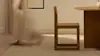 A person in a sheer dress walks barefoot past a wooden chair and round wooden table in a softly lit, minimalist room.