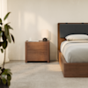 A modern bedroom with a wooden bed frame and matching nightstand, featuring a lamp, a book, and a cup. A plant is visible in the foreground, and soft natural light fills the space.