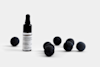 A small glass dropper bottle labeled Deep Forest Parfum DAmbiance stands on a white surface beside several scattered dark spherical objects. The background is plain and light gray.