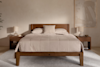 A neatly made bed with beige bedding and three pillows stands between two wooden nightstands, each holding a lamp and decor, against a neutral wall with abstract art. A light rug covers the floor.