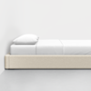 A minimalistic beige bed with a white pillow and white bedding, positioned against a plain white wall on a light gray floor. The scene is clean and modern with a simple aesthetic.