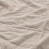 A close-up view of a soft, quilted beige fabric with visible stitching forming a square pattern, showing gentle folds and a slightly wrinkled texture.
