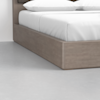 Close-up view of a corner of a bed with a wooden frame and white bedding, including two pillows, on a smooth light gray floor and against a white wall.