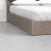 Close-up view of a corner of a bed with a wooden frame and white bedding, including two pillows, on a smooth light gray floor and against a white wall.