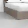 Close-up view of a corner of a bed with a wooden frame and white bedding, including two pillows, on a smooth light gray floor and against a white wall.