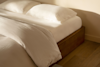 A neatly made bed with white sheets, pillows, and a comforter sits on a wooden frame in a sunlit room with light-colored walls and wood flooring.