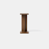 A small, modern wooden pedestal table with a square top and base, featuring a simple, minimalist design against a plain white background.