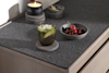 A glass of iced drink with lime slices sits on a coaster next to a bowl of cherries, a lit candle, and a small stone container with wooden sticks on a grey countertop.