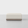 A modern, rectangular beige ottoman with a textured fabric top and a smooth wooden base, set on a light gray floor against a plain white background.