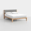 A modern bed with a light wood frame and legs, featuring a gray upholstered headboard. The bed is made with white sheets and two white pillows, set against a plain white wall and floor.
