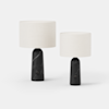 Two modern table lamps with cylindrical white fabric shades and black marble bases are displayed side by side on a plain white background. The bases have a rounded design and a small circular switch.