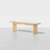 A minimalist wooden bench with a light-colored rectangular seat and two wide, flat legs, positioned on a smooth, light gray floor against a plain white background.