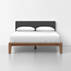 A modern bed with a dark gray upholstered headboard, wooden frame, and white bedding, including two white pillows, positioned against a plain white wall on a light gray floor.