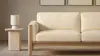 A beige fabric sofa with wooden frame sits next to a stone side table with a small white lamp on it. Two framed artworks hang on the beige wall in the background.