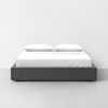 A modern, low-profile bed with a dark upholstered frame, white bedding, and two pillows, set against a plain white wall and light gray floor.