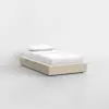 A minimalist single bed with a beige upholstered base, white bedding, and a pillow, set against a plain white background in a modern, empty room.